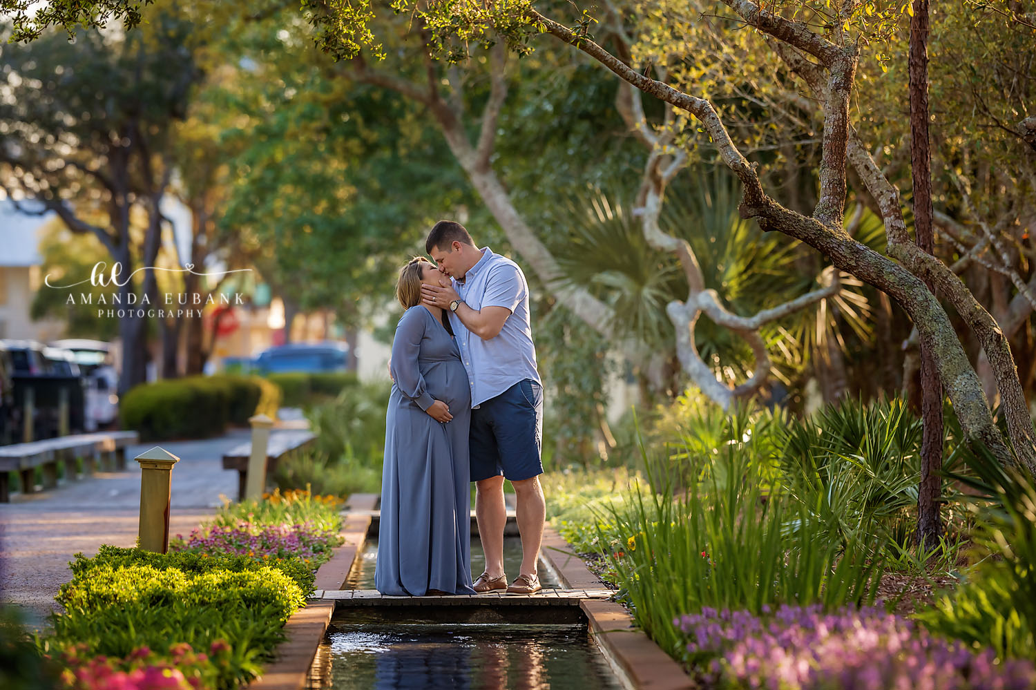 Expecting couple standing by a landscaped garden fountain, the man kissing the pregnant woman’s forehead as she wears a long blue dress, photographed by a Santa Rosa Beach maternity photographer