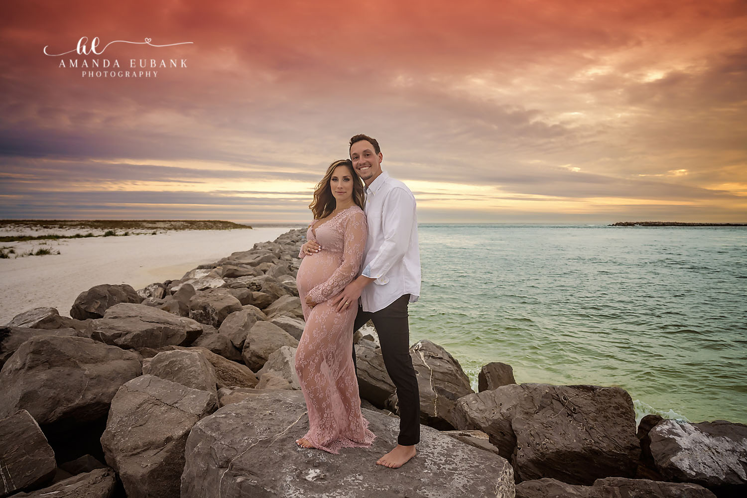 Santa Rosa Beach Maternity Photographer – Sunset Coastal Rocks Maternity Portrait Expecting couple standing on coastal rocks at sunset, the pregnant woman in a pink lace maternity gown cradling her belly beside the man in a white shirt with the ocean behind, photographed by a Santa Rosa Beach maternity photographer
