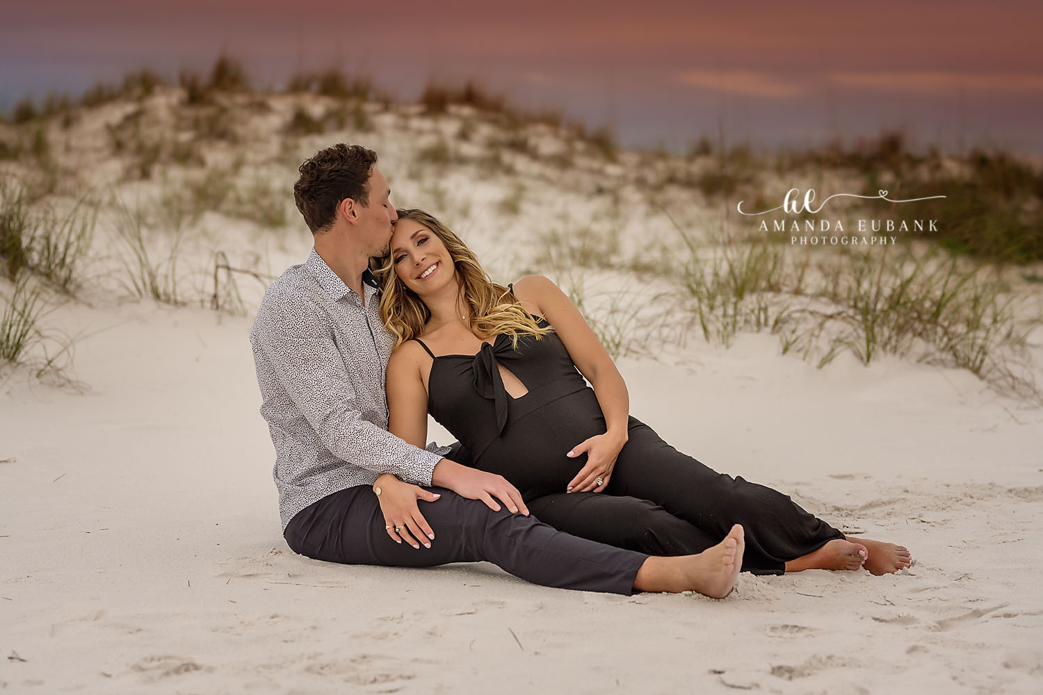 Expecting couple sitting on the beach sand near the dunes, the pregnant woman in a black maternity jumpsuit cradling her belly as the man kisses her temple, photographed by a Santa Rosa Beach maternity photographer
