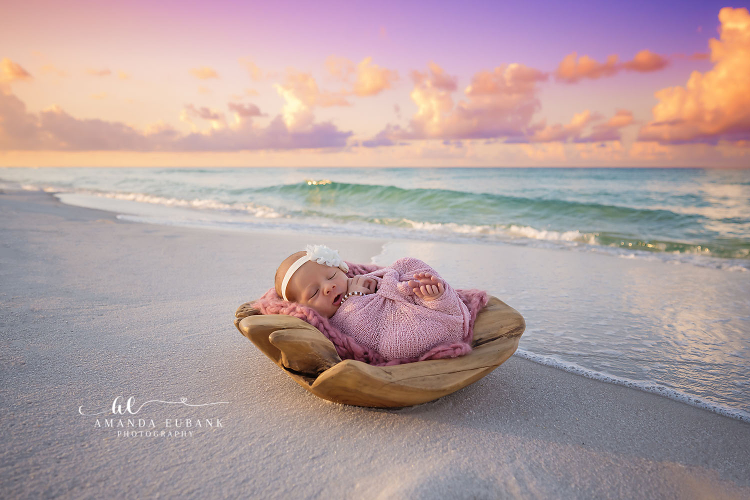 Newborn baby girl in a lavender swaddle with a floral headband sleeping in a wooden bowl on the beach near the shoreline at sunset, photographed by a Santa Rosa Beach newborn photographer