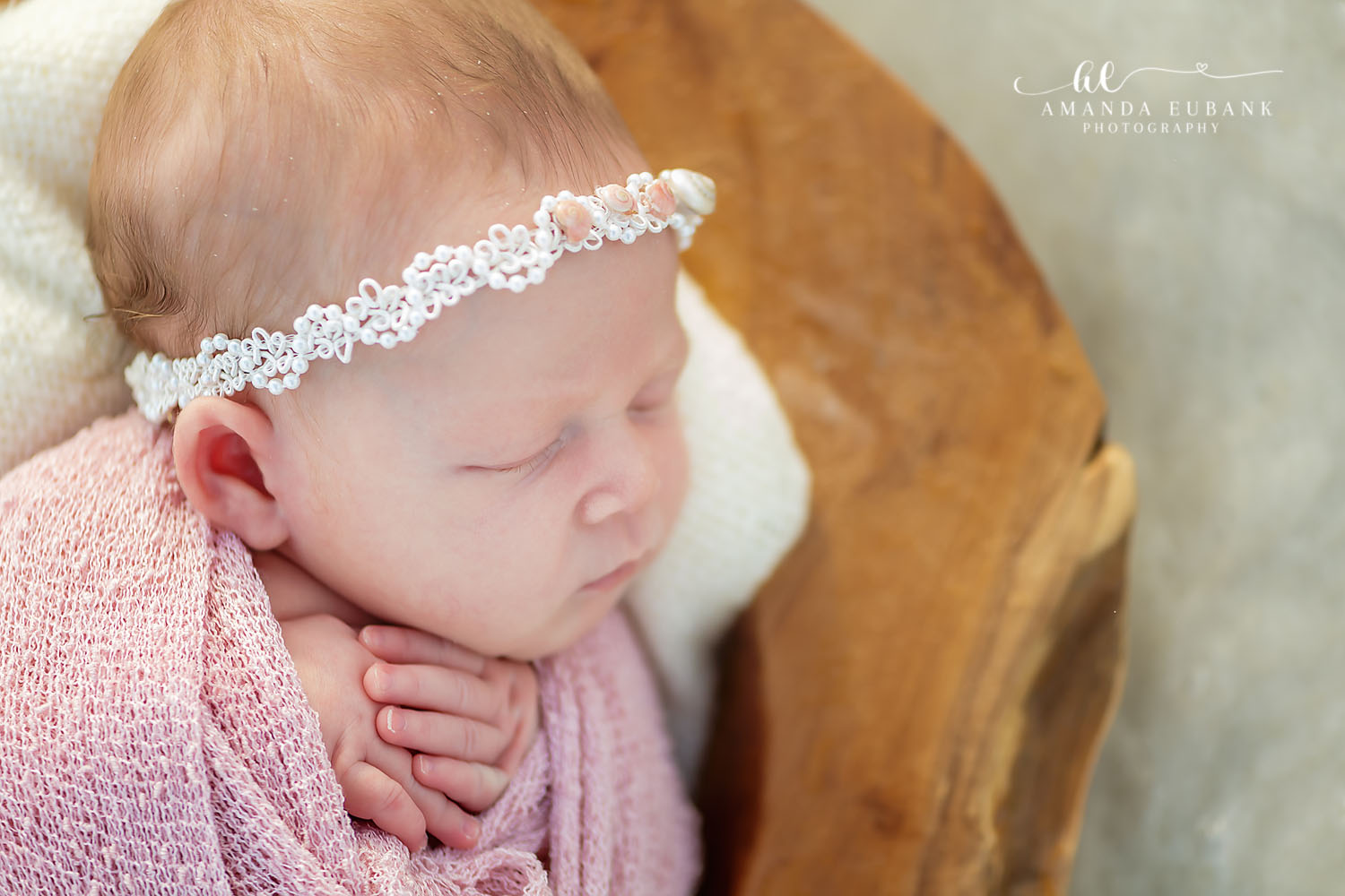 Sleeping newborn baby girl wrapped in a soft pink swaddle with a white lace headband, resting against a wooden prop, photographed by a Santa Rosa Beach newborn photographer