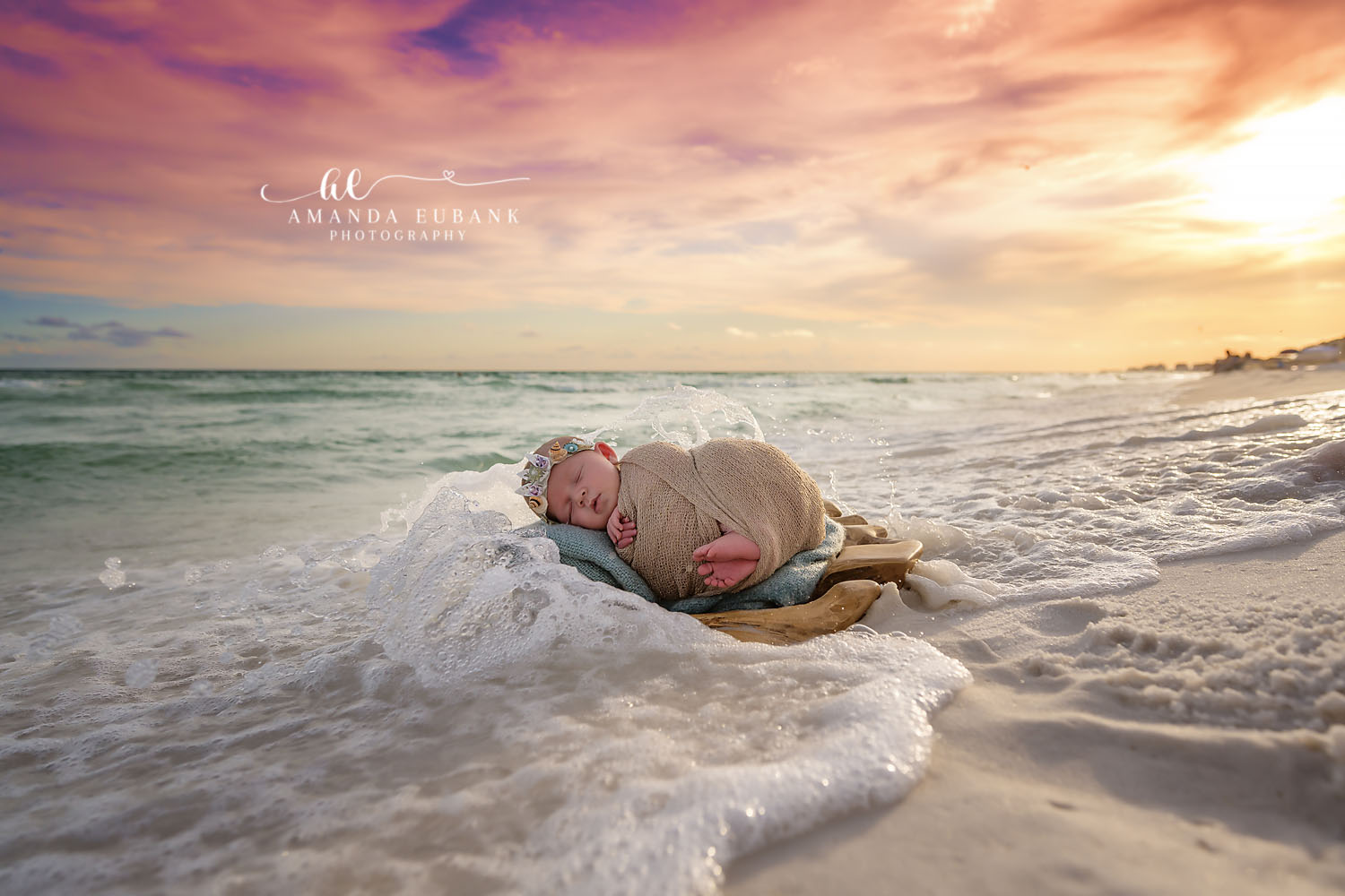 Santa Rosa Beach Newborn Photographer – Sunset Newborn Portrait at the Water’s Edge Newborn baby wrapped in a tan swaddle with a floral headband sleeping on driftwood at the water’s edge as waves wash around at sunset, photographed by a Santa Rosa Beach newborn photographer