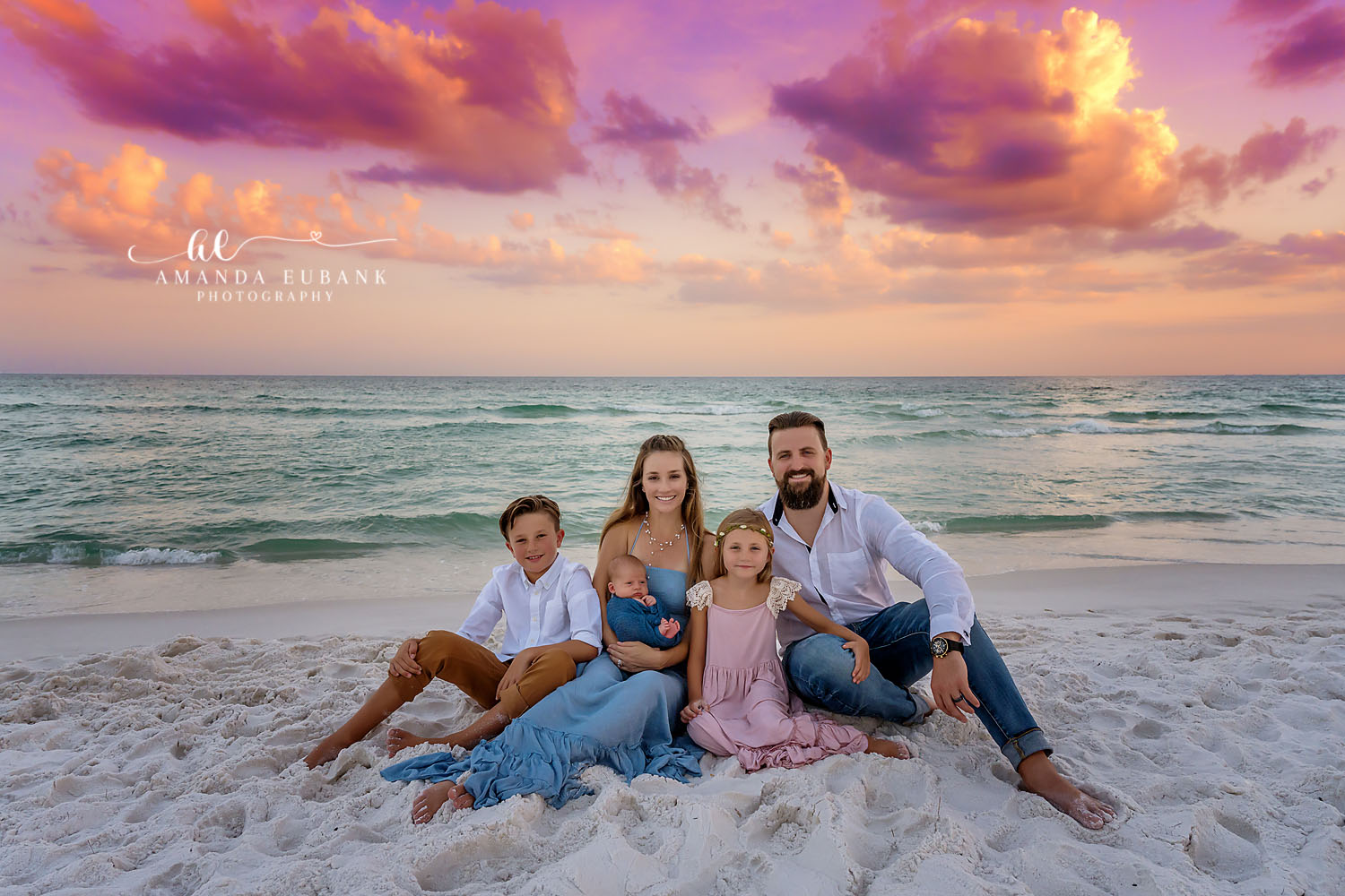 Family of five on the beach at sunset, the mother in a blue dress holding a newborn baby beside the father and two older children, photographed by a Santa Rosa Beach newborn photographer