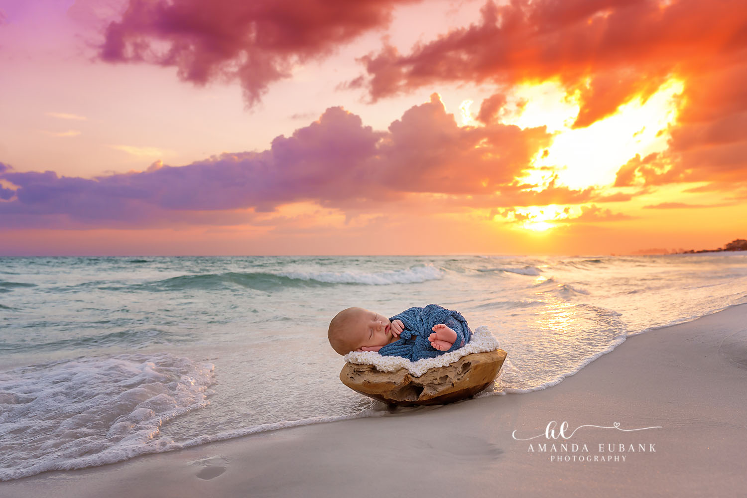 Newborn baby in a blue outfit sleeping in a wooden bowl on the sand at sunset, photographed by a Santa Rosa Beach newborn photographer