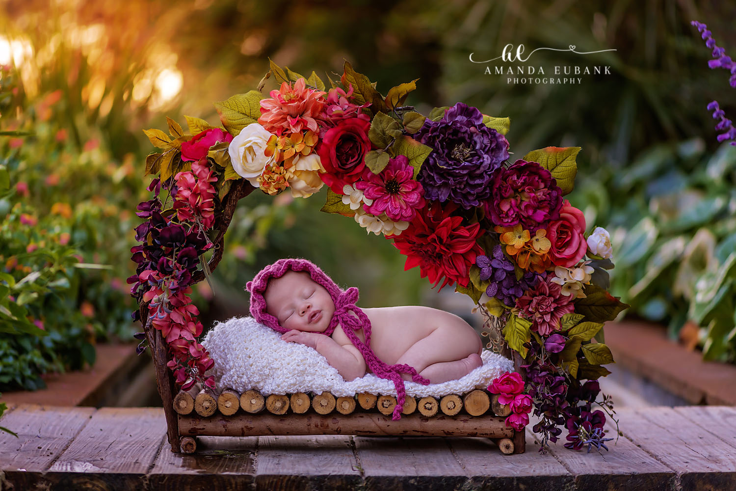 Santa Rosa Beach Newborn Photographer – Floral Garden Newborn Portrait Newborn baby in a pink bonnet sleeping in a wooden bed surrounded by a colorful floral arch in a garden, taken by a Santa Rosa Beach newborn photographer