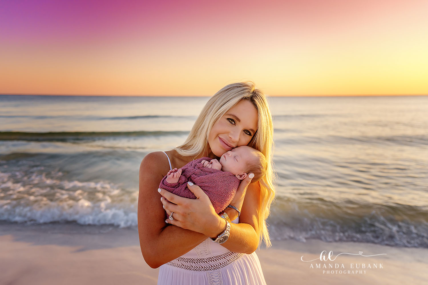 A smiling mother holding her newborn baby close to her cheek on the beach at sunset, taken by a Santa Rosa Beach newborn photographer