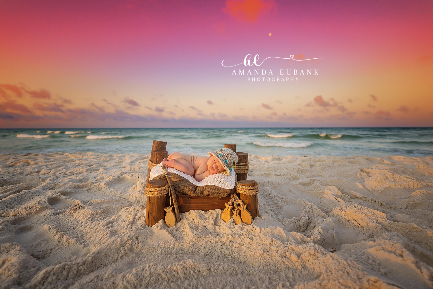 Newborn baby sleeping on a small wooden bed with nautical decor on the beach under a vivid pink and purple sunset, taken by a Santa Rosa Beach newborn photographer