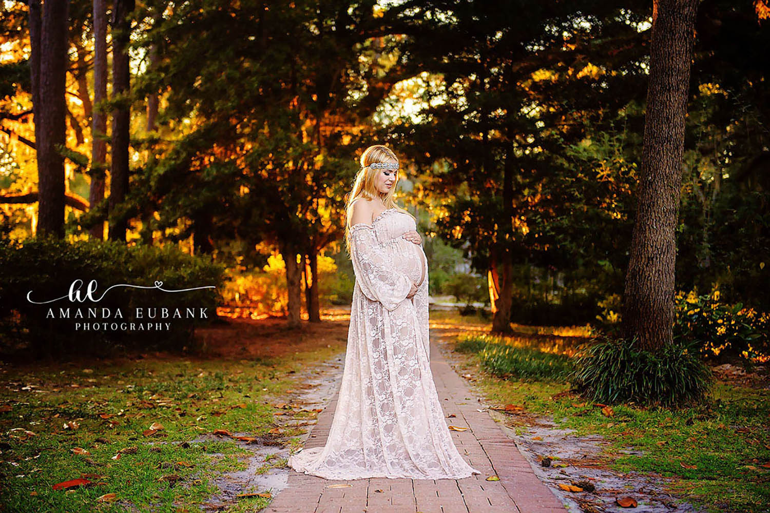 Maternity portrait of an expecting woman in a flowing cream lace gown cradling her belly on a brick path in a wooded park at golden hour, taken by a Santa Rosa Beach maternity photographer