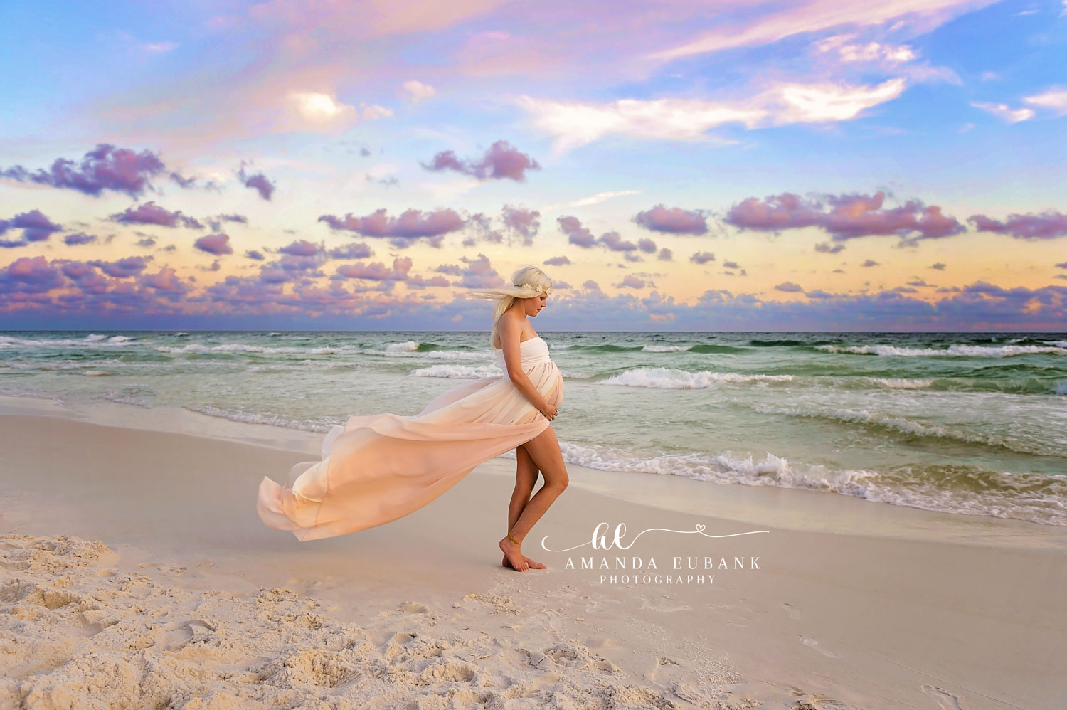 Maternity portrait of an expecting woman in a flowing peach gown walking along the shoreline at sunset as the fabric catches the wind, taken by a Santa Rosa Beach maternity photographer