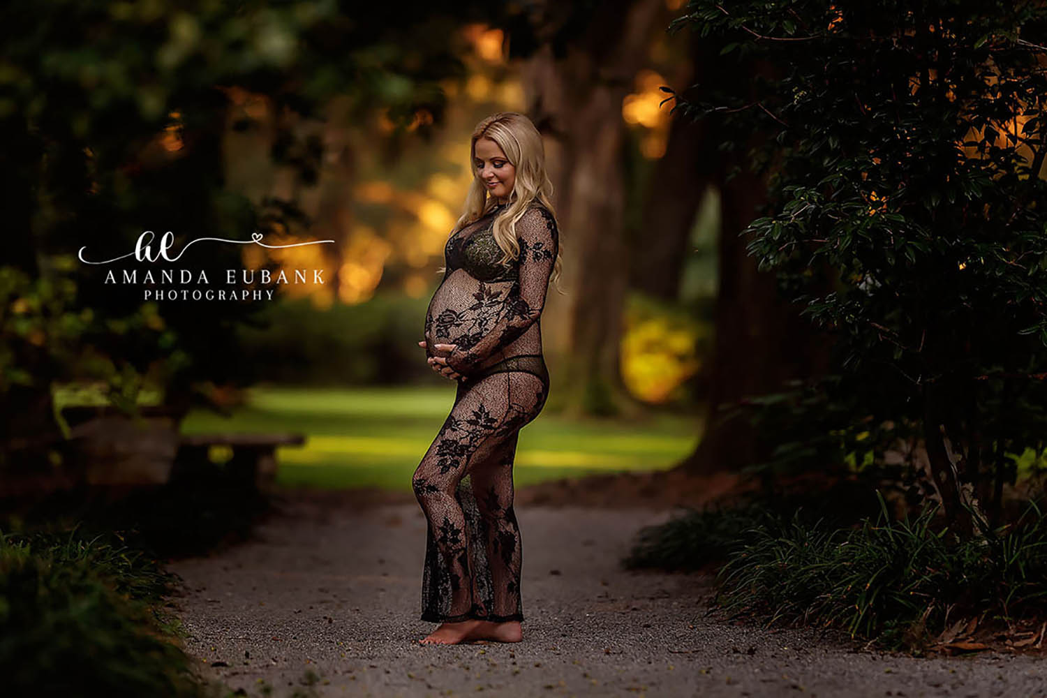 Santa Rosa Beach Photographer Maternity portrait of an expecting woman in a black lace gown cradling her belly on a wooded path in warm golden light, taken by a Santa Rosa Beach maternity photographer