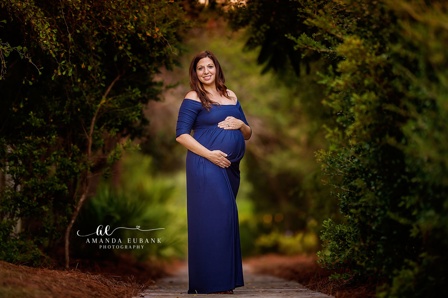 Maternity portrait of an expecting woman in a navy off-the-shoulder gown cradling her belly on a path framed by a green tree archway, taken by a Santa Rosa Beach maternity photographer