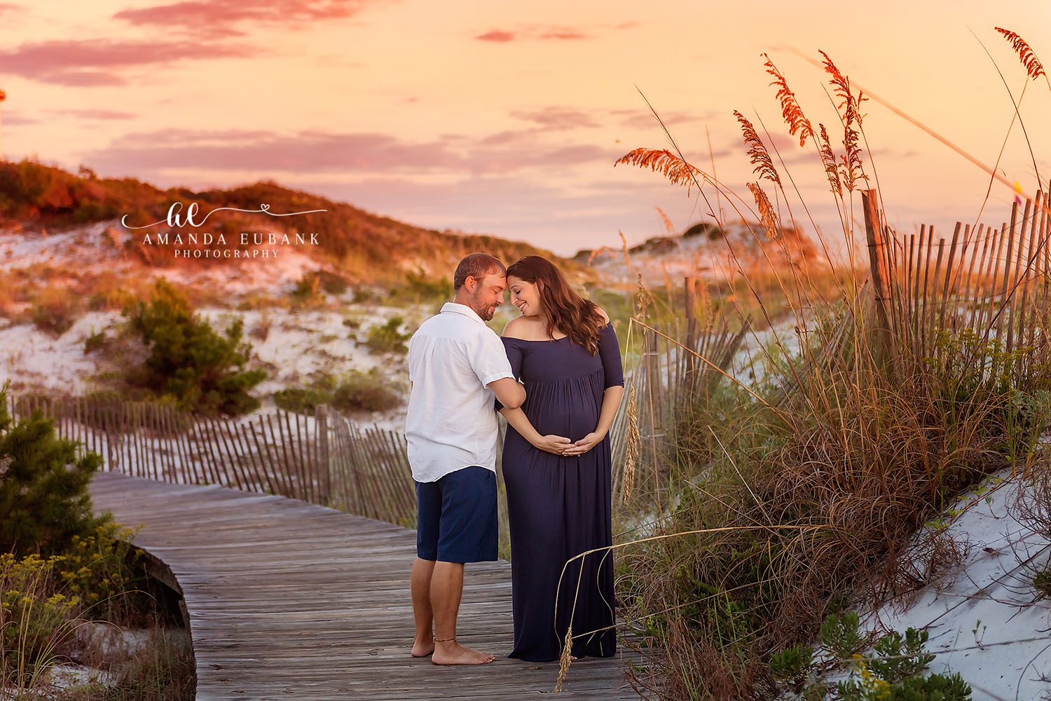 Maternity portrait of an expecting couple standing on a dune boardwalk among sea oats at sunset with the woman in a navy gown, taken by a Santa Rosa Beach maternity photographer