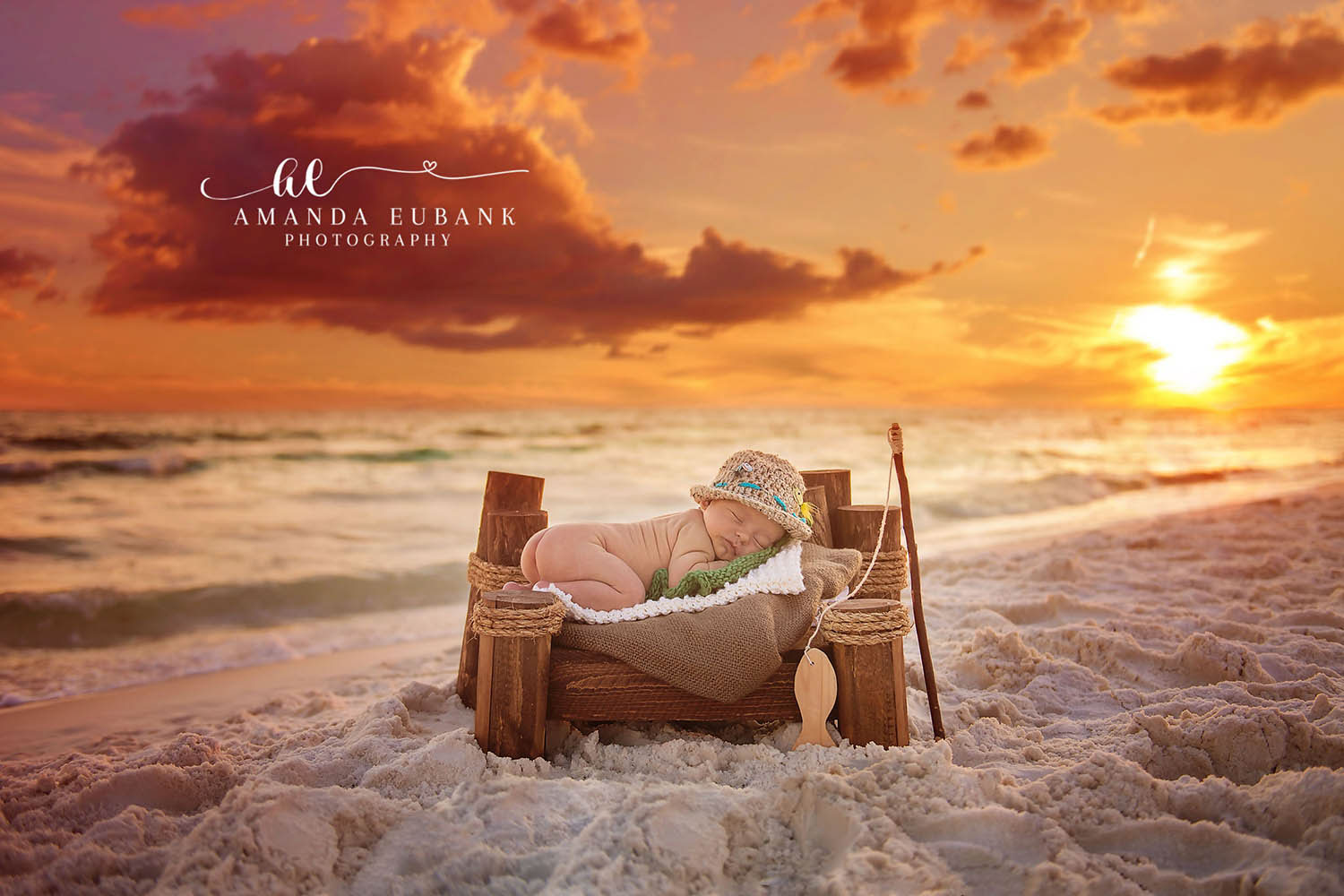 Newborn baby in a fisherman-themed outfit with a fishing pole sleeping on a wooden bed on the beach at golden sunset, taken by a Santa Rosa Beach newborn photographer