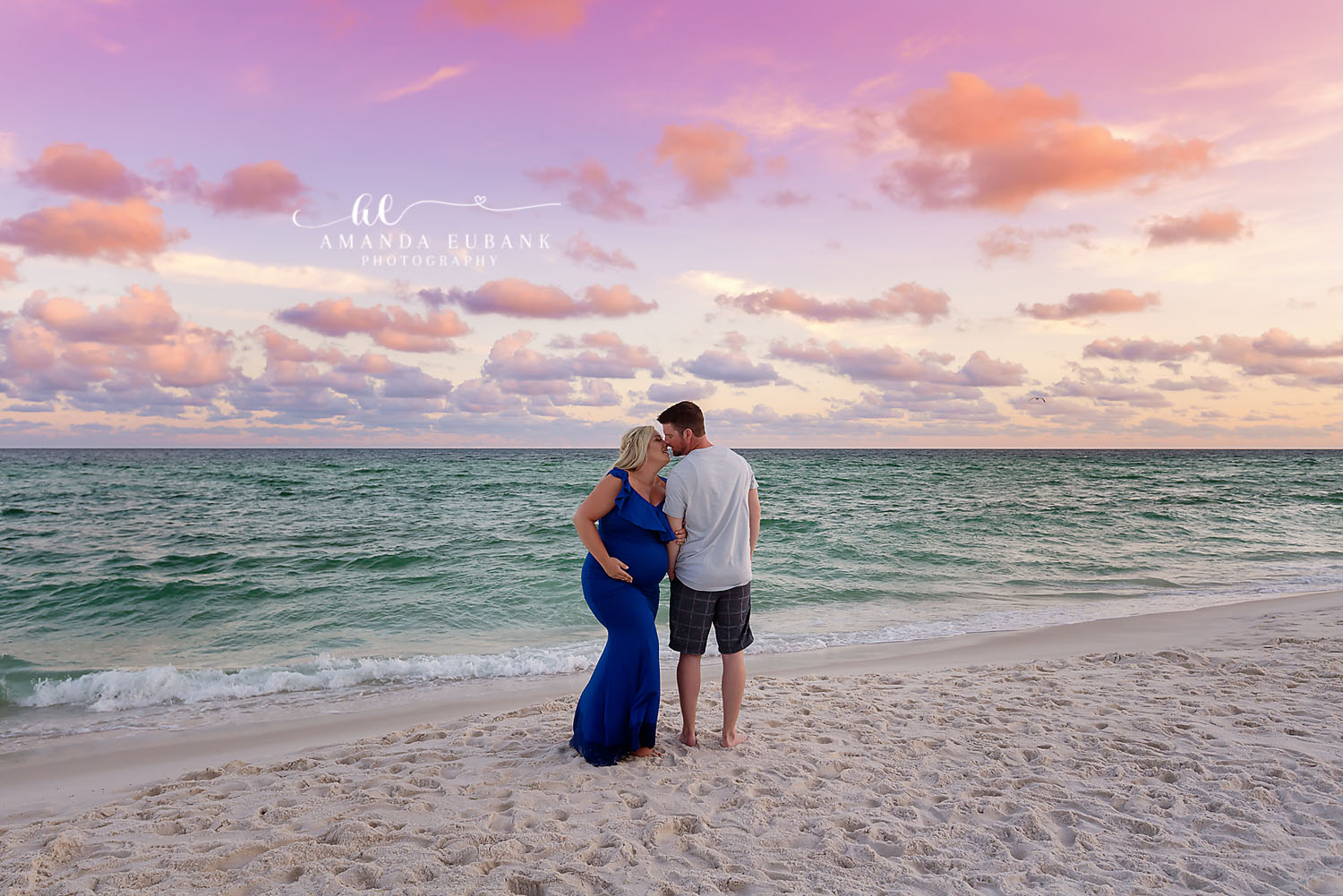 Maternity portrait of an expecting couple embracing at the shoreline with the woman in a navy gown under a pastel sunset sky, taken by a Santa Rosa Beach maternity photographer