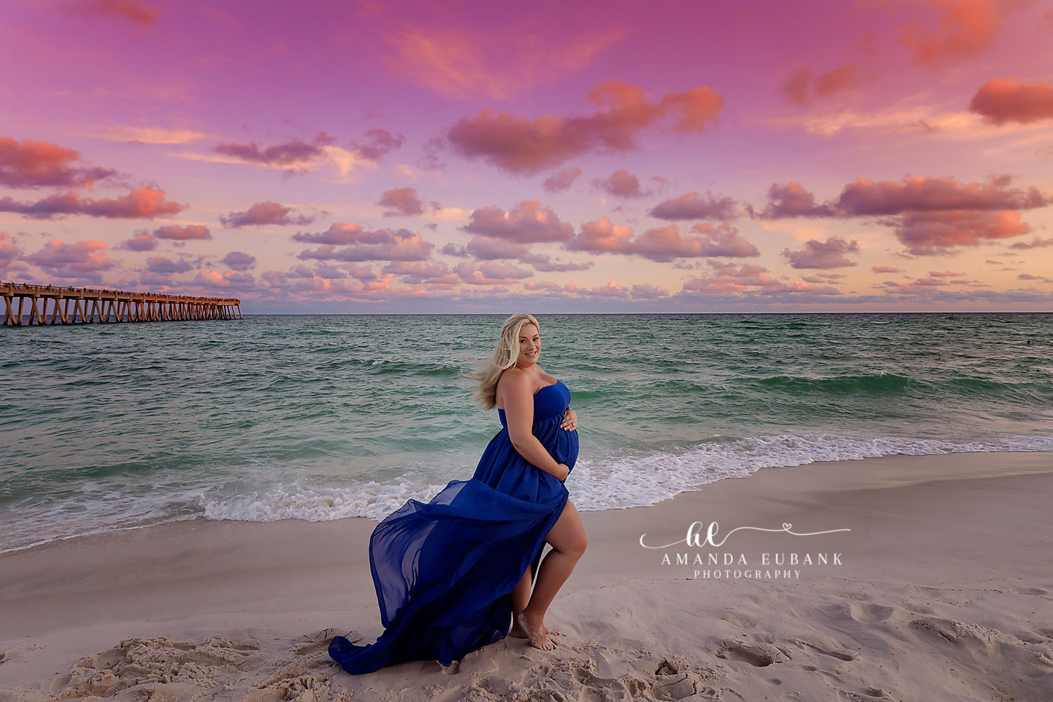 Santa Rosa Beach Photographer Maternity portrait of an expecting woman in a flowing royal blue gown posing on the beach at sunset near a pier, taken by a Santa Rosa Beach maternity photographer