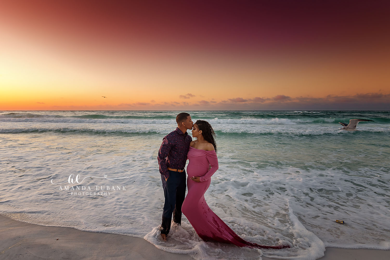Maternity portrait of an expecting couple at the shoreline at sunset with the woman in a flowing pink gown, taken by a Santa Rosa Beach maternity photographer