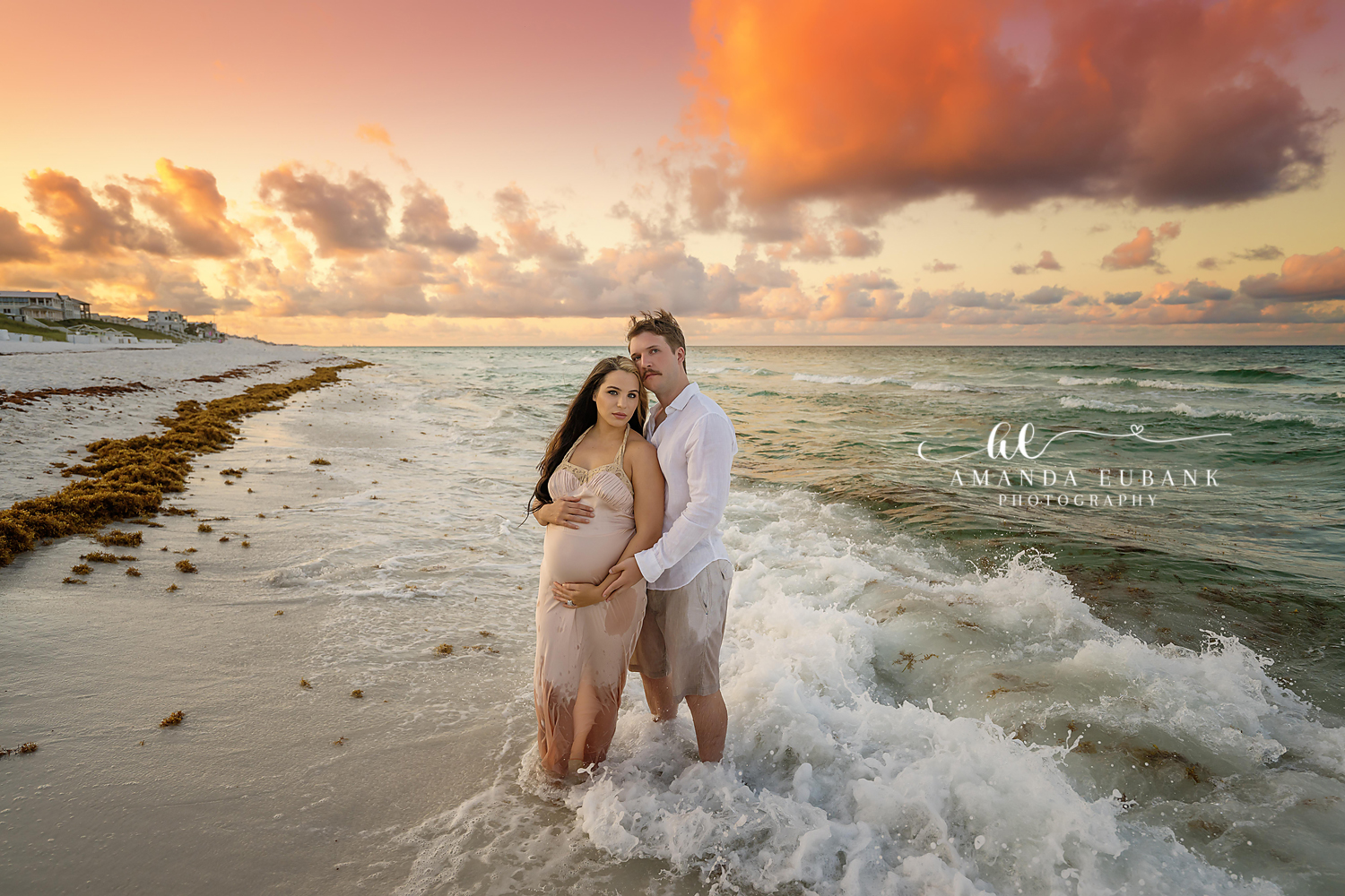 Maternity portrait of an expecting couple standing in the shoreline waves at sunset as she cradles her belly, taken by a Santa Rosa Beach maternity photographer