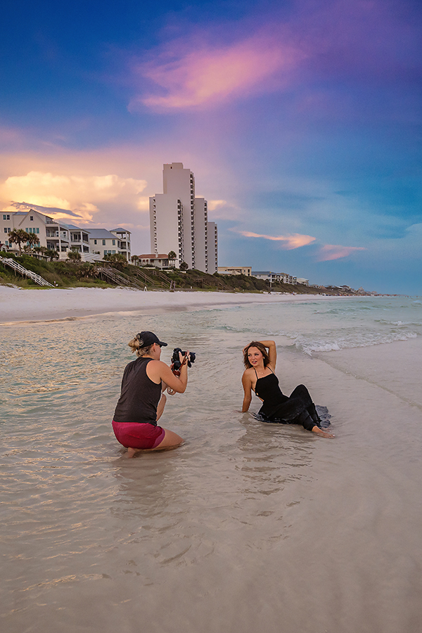 Santa Rosa Beach Photographer – Behind the Scenes Beach Portrait Session Behind-the-scenes photo of a Santa Rosa Beach photographer with a camera photographing a woman posing in the shallow water at sunset along the coastline