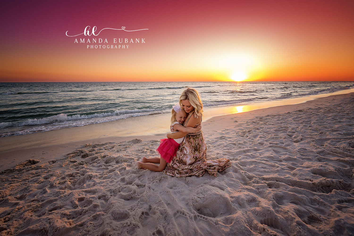 Santa Rosa Beach Family Photographer – Mother and Daughter at Sunset Mother and young daughter sharing a tender embrace while kneeling on the sand at sunset, the mother in a patterned dress and the little girl in a pink tutu, with a golden sunset over the ocean, photographed by a Santa Rosa Beach family photographer