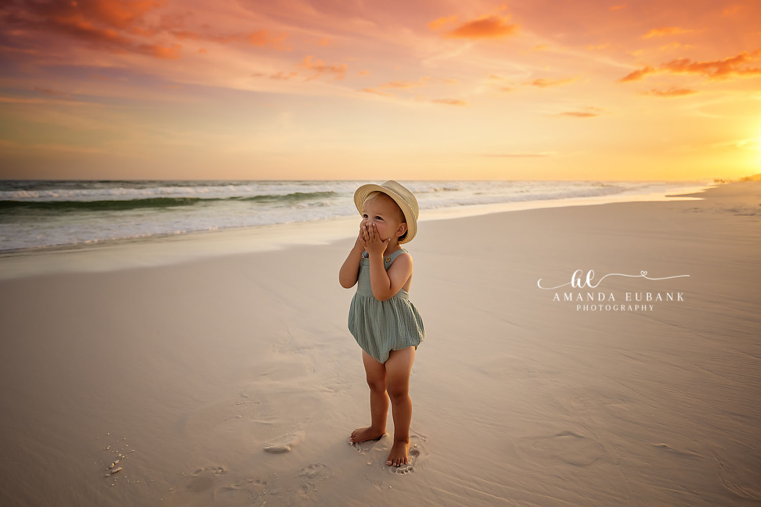 Toddler in a sage-green romper and straw sun hat standing on the sand at sunset with hands to her face in a sweet gesture, photographed by a Santa Rosa Beach family photographer