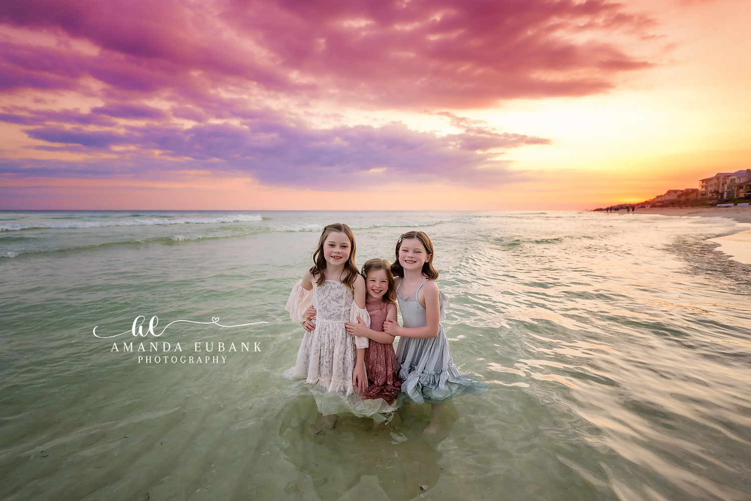 Three young sisters standing together in the shallow ocean at sunset wearing flowing pastel dresses under a soft pink sky, photographed by a Santa Rosa Beach family photographer