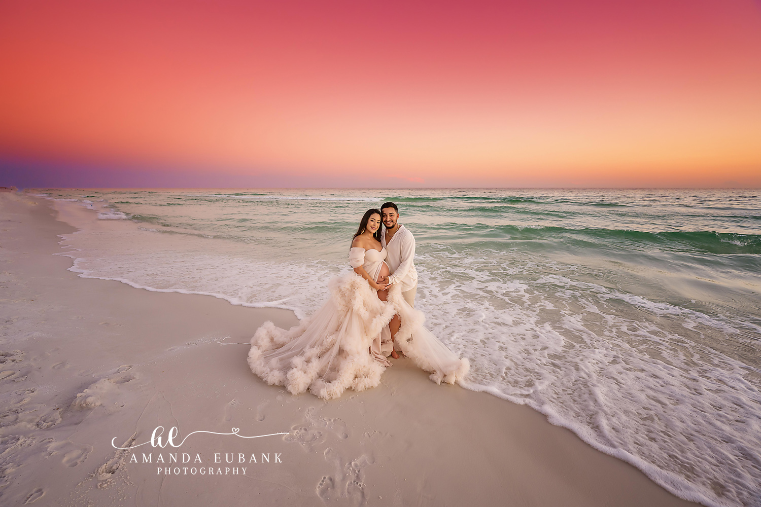 Expecting couple kneeling together on the beach at sunset, the pregnant woman in a flowing cream tulle gown cradling her belly beside her partner in white, photographed by a Santa Rosa Beach maternity photographer