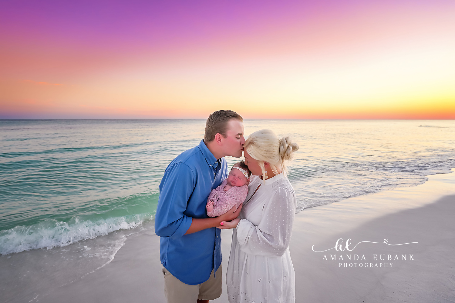 Santa Rosa Beach Newborn Photographer – New Parents with Newborn at Sunset New parents on the beach at sunset, the father in blue cradling their newborn baby and kissing the mother in white on the forehead, photographed by a Santa Rosa Beach newborn photographer