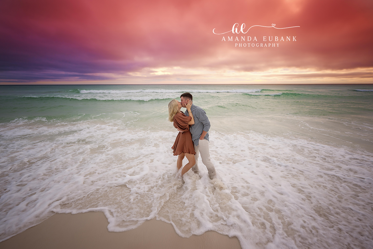 Couple kissing in the surf at sunset, the woman in a rust dress and the man in a chambray shirt as waves wash around their feet, photographed by a Santa Rosa Beach engagement photographer