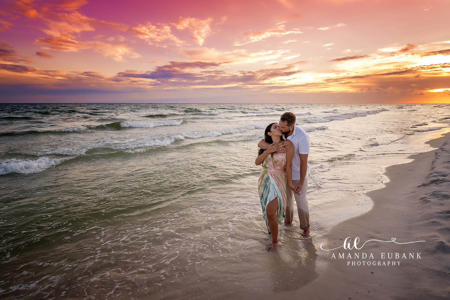 Couple embracing at the water’s edge at sunset, the man kissing the woman’s forehead as she wears a flowing floral dress along the shoreline, photographed by a Santa Rosa Beach engagement photographer