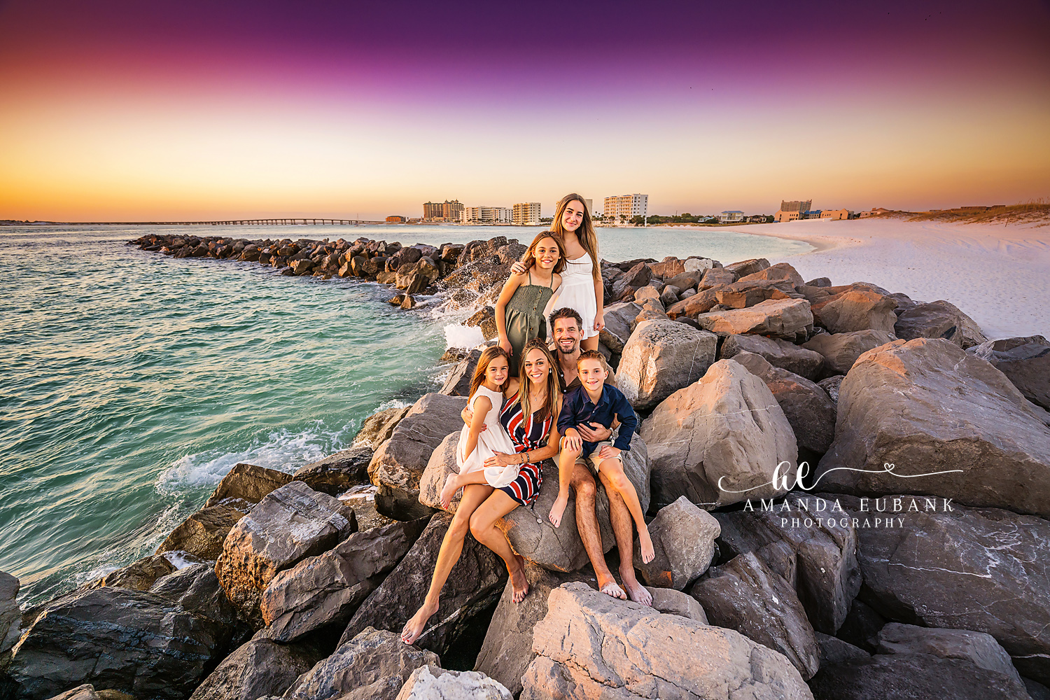 Family of six posed together on a rocky jetty by the water at sunset with the coastline in the distance, photographed by a Santa Rosa Beach family photographer