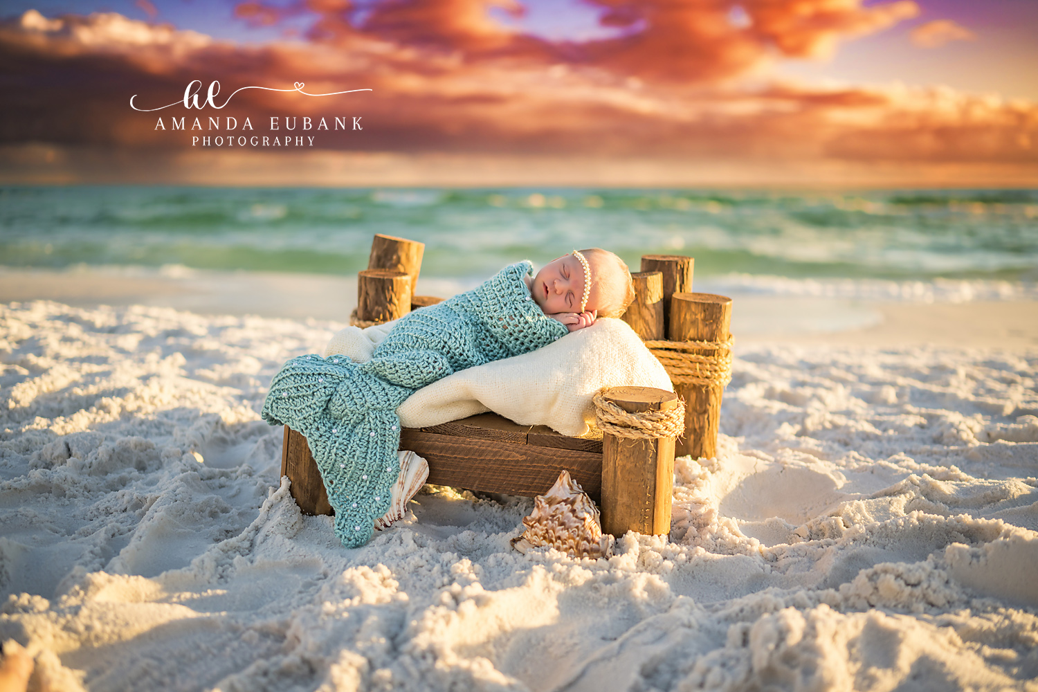 Sleeping newborn baby in a teal knit mermaid-tail outfit posed on a wooden bed prop with a seashell on the white sand beach, photographed by a Santa Rosa Beach newborn photographer