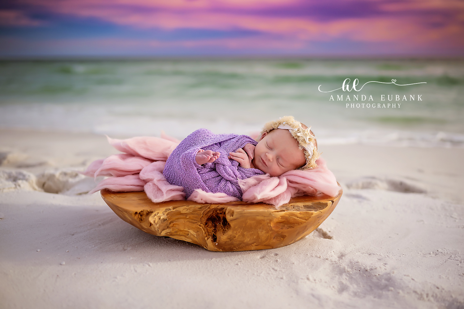 Sleeping newborn baby wrapped in a purple swaddle with a lace headband, nestled in a wooden bowl with soft pink fabric on the white sand beach, photographed by a Santa Rosa Beach newborn photographer
