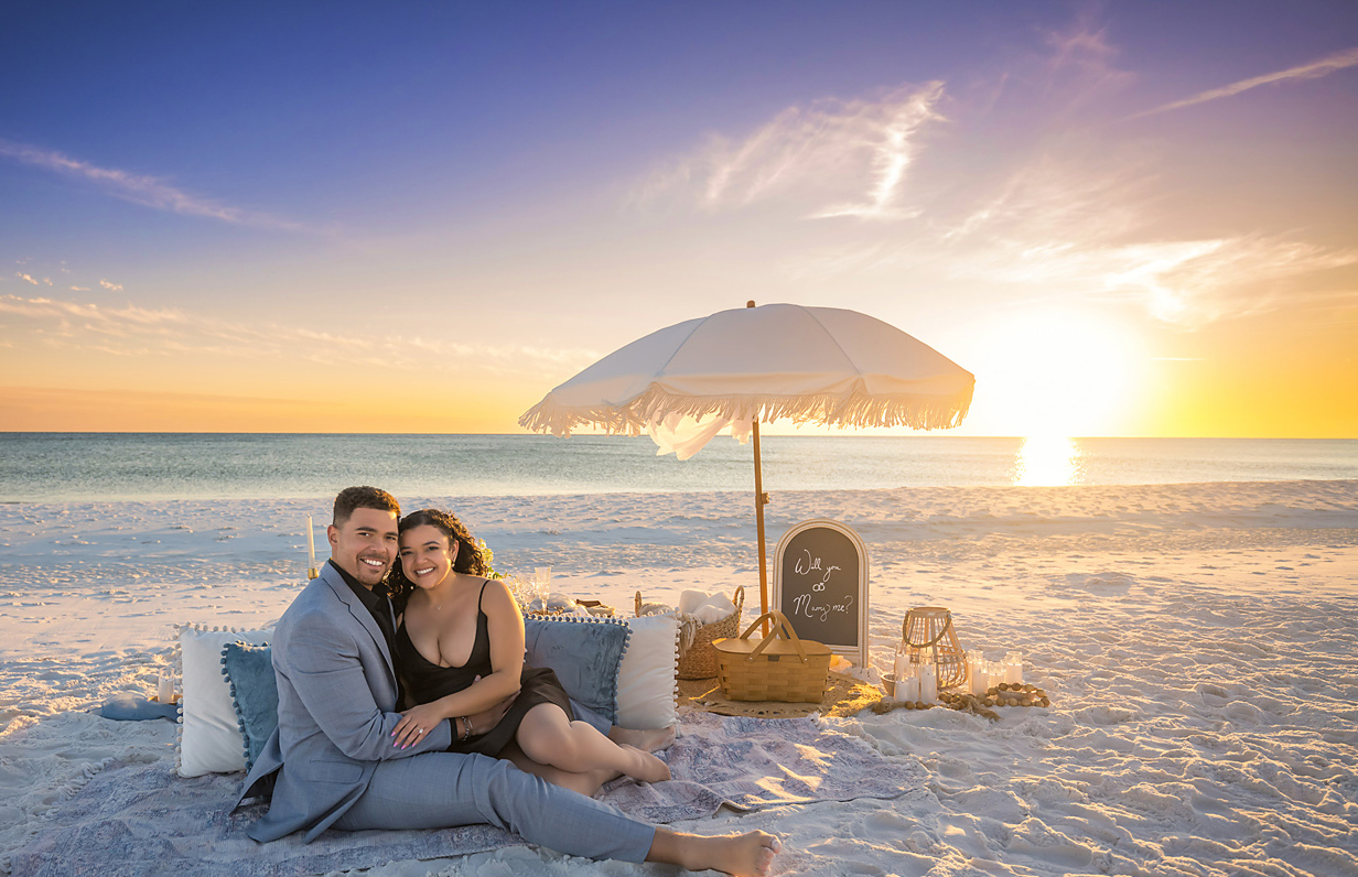 Couple cuddled together on a styled beach picnic setup at sunset with a fringed umbrella, pillows, candles and a Will You Marry Me chalkboard sign, photographed by a Santa Rosa Beach engagement photographer