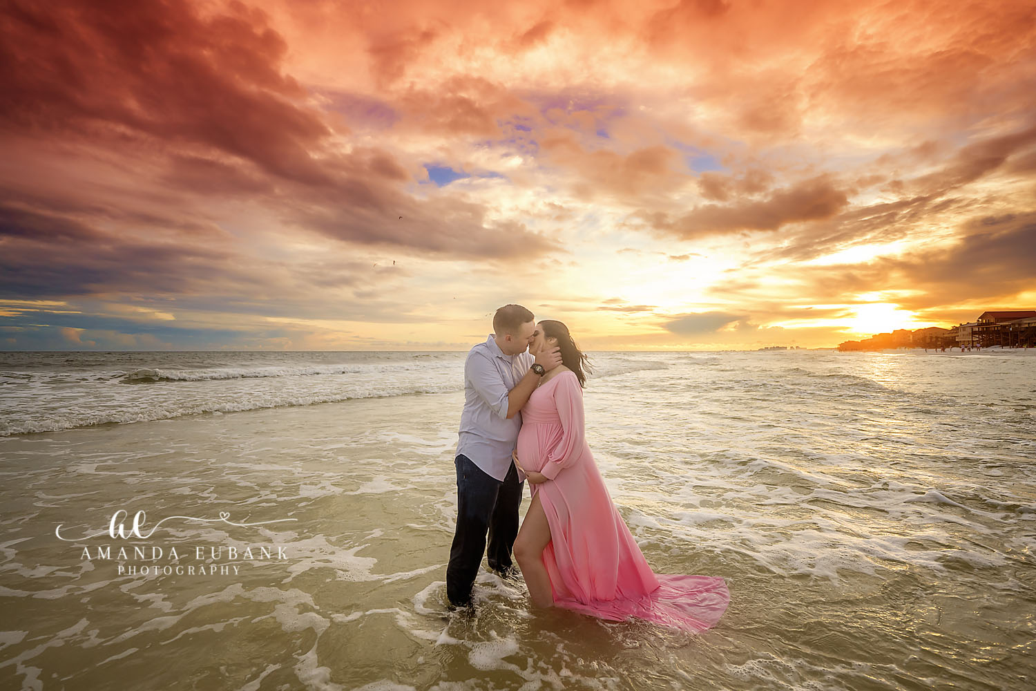 Expecting couple sharing a kiss in the surf at sunset, the woman in a flowing pink maternity gown as her partner cradles her belly, photographed by a Santa Rosa Beach maternity photographer