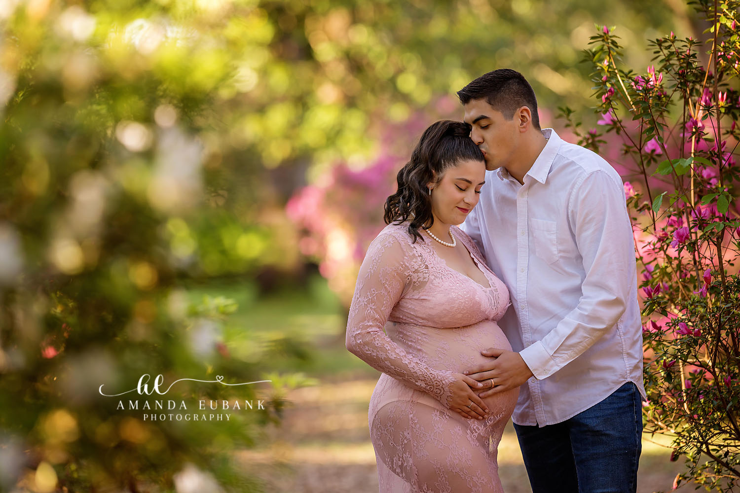 Expecting couple in a flowering garden, the woman in a pink lace maternity gown cradling her belly while her partner kisses her head, photographed by a Santa Rosa Beach maternity photographer