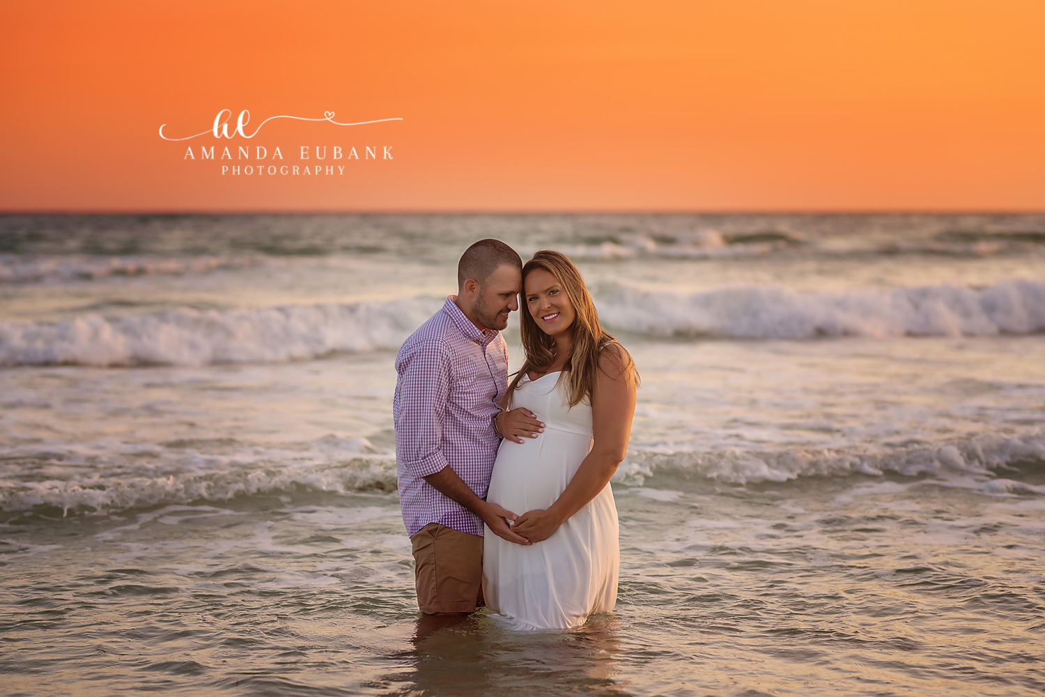 Expecting couple standing together in the surf at sunset, the woman in a white maternity dress as both cradle her belly, photographed by a Santa Rosa Beach maternity photographer