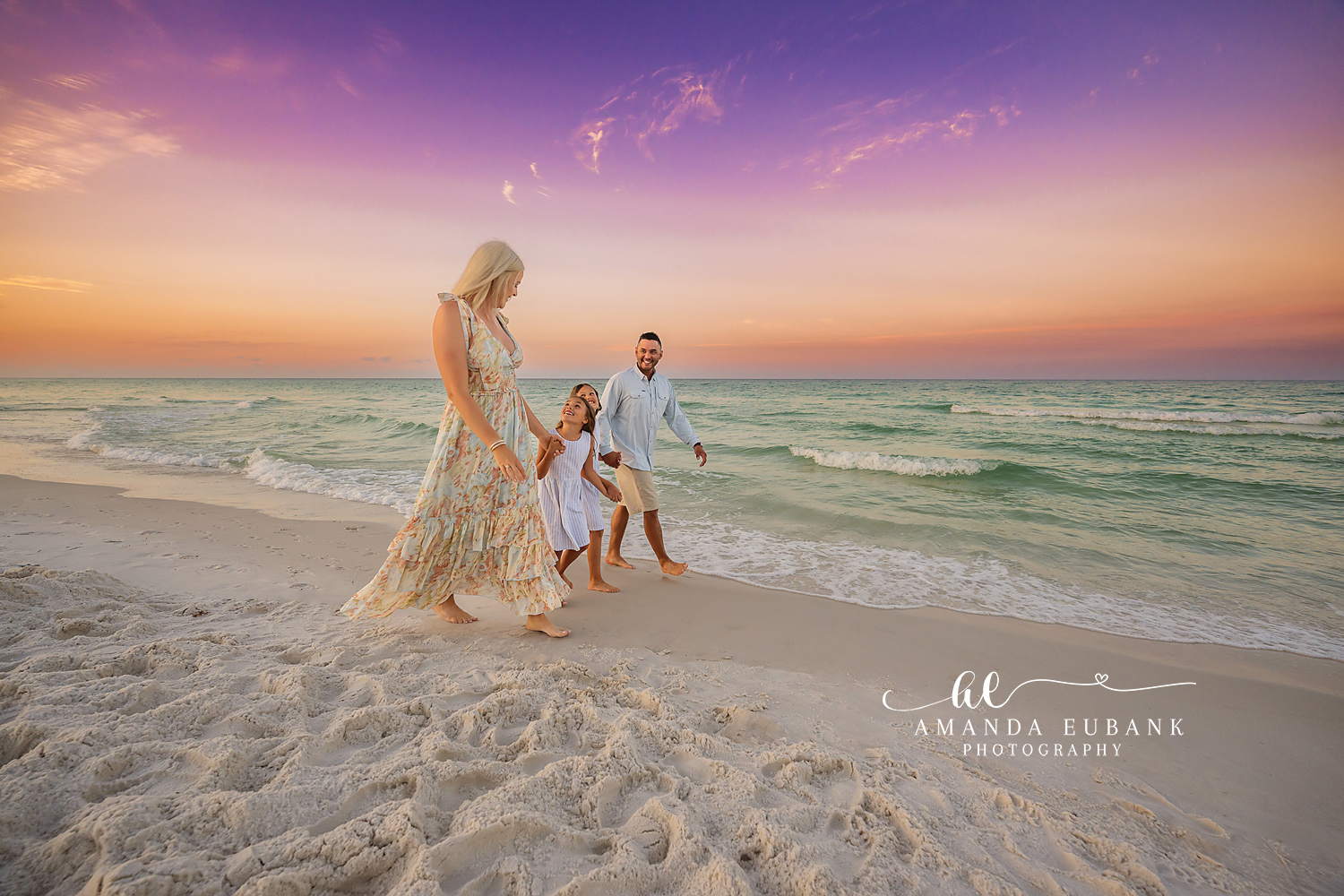 Family of three walking hand in hand along the shoreline at sunset, the mother in a floral dress and child in white between her parents, photographed by a Santa Rosa Beach family photographer