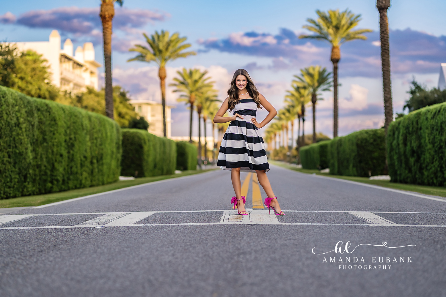 Young woman in a black and white striped dress and pink heels posing in the middle of a manicured hedge-lined avenue, photographed by a Santa Rosa Beach senior photographer