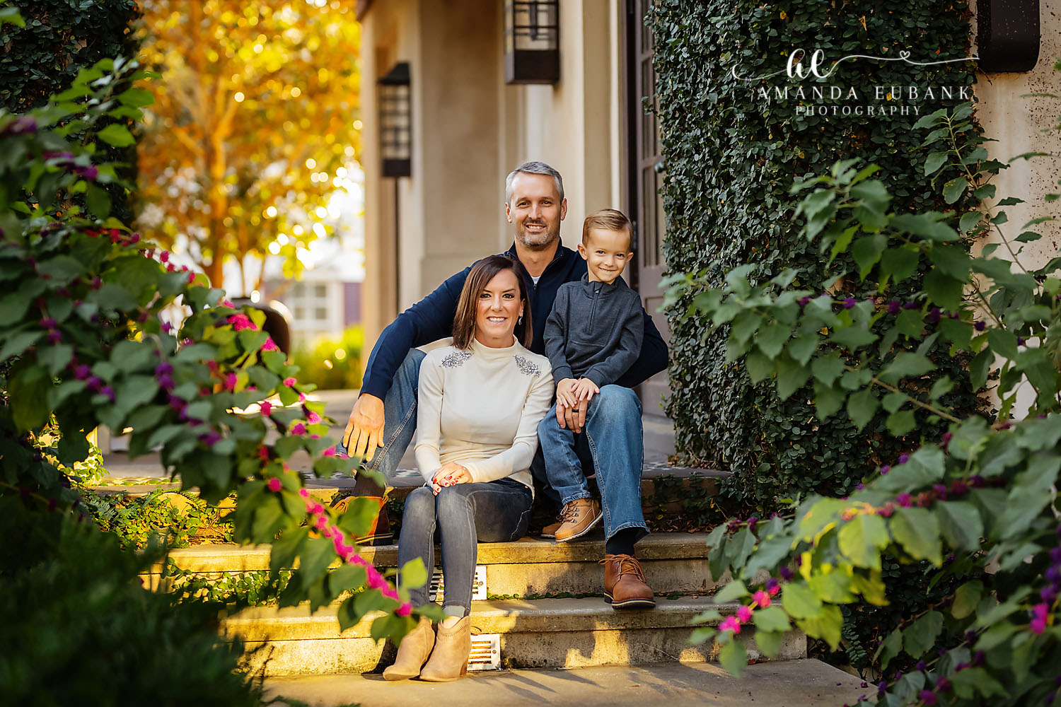 Santa Rosa Beach Family Photographer – Family Portrait on Garden Steps Family of three sitting together on stone steps framed by lush greenery and pink blooms during a fall portrait session, photographed by a Santa Rosa Beach family photographer