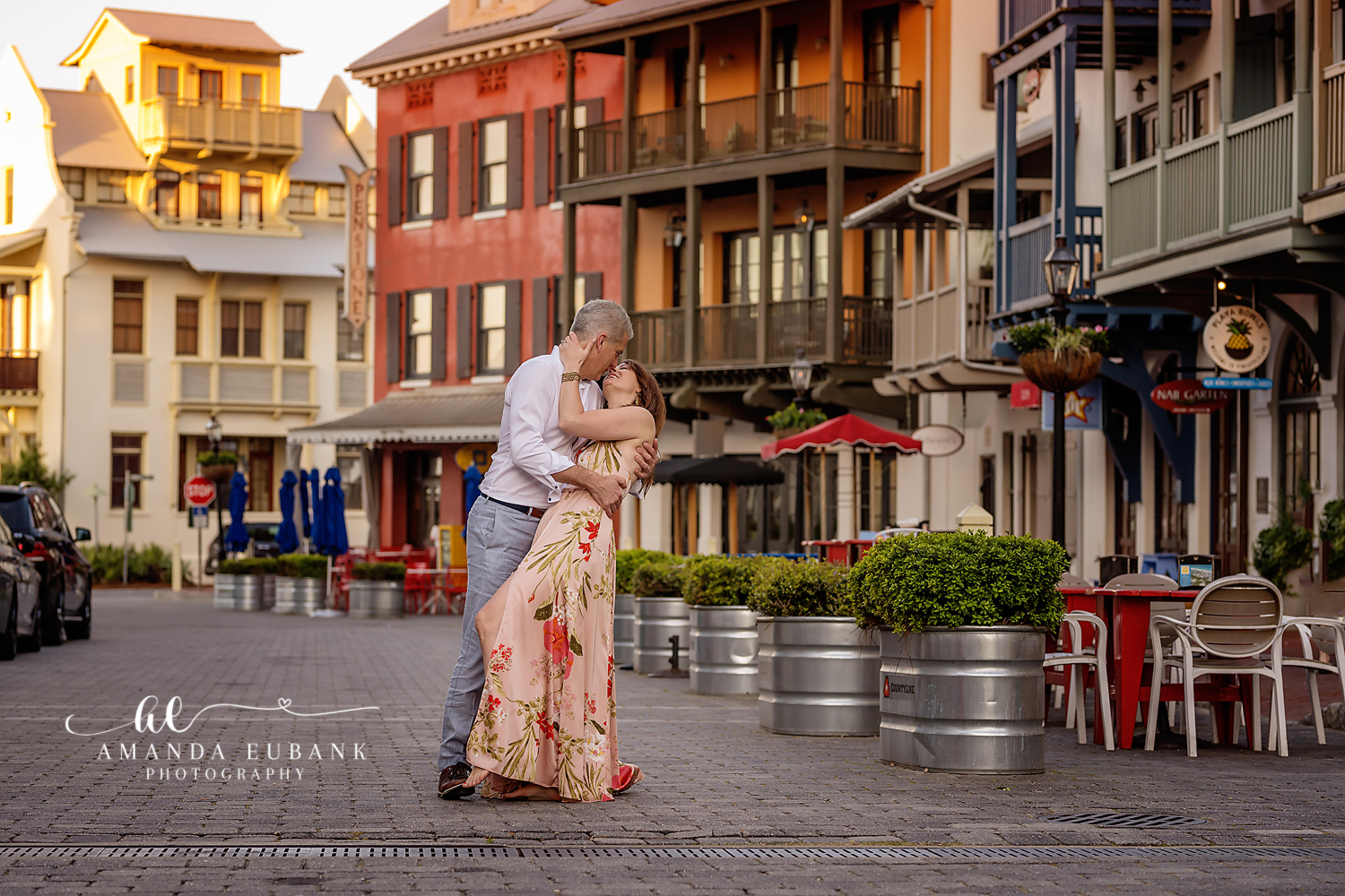 Couple sharing a romantic dip embrace in a charming town square lined with cafes, the woman in a floral gown, photographed by a Santa Rosa Beach engagement photographer