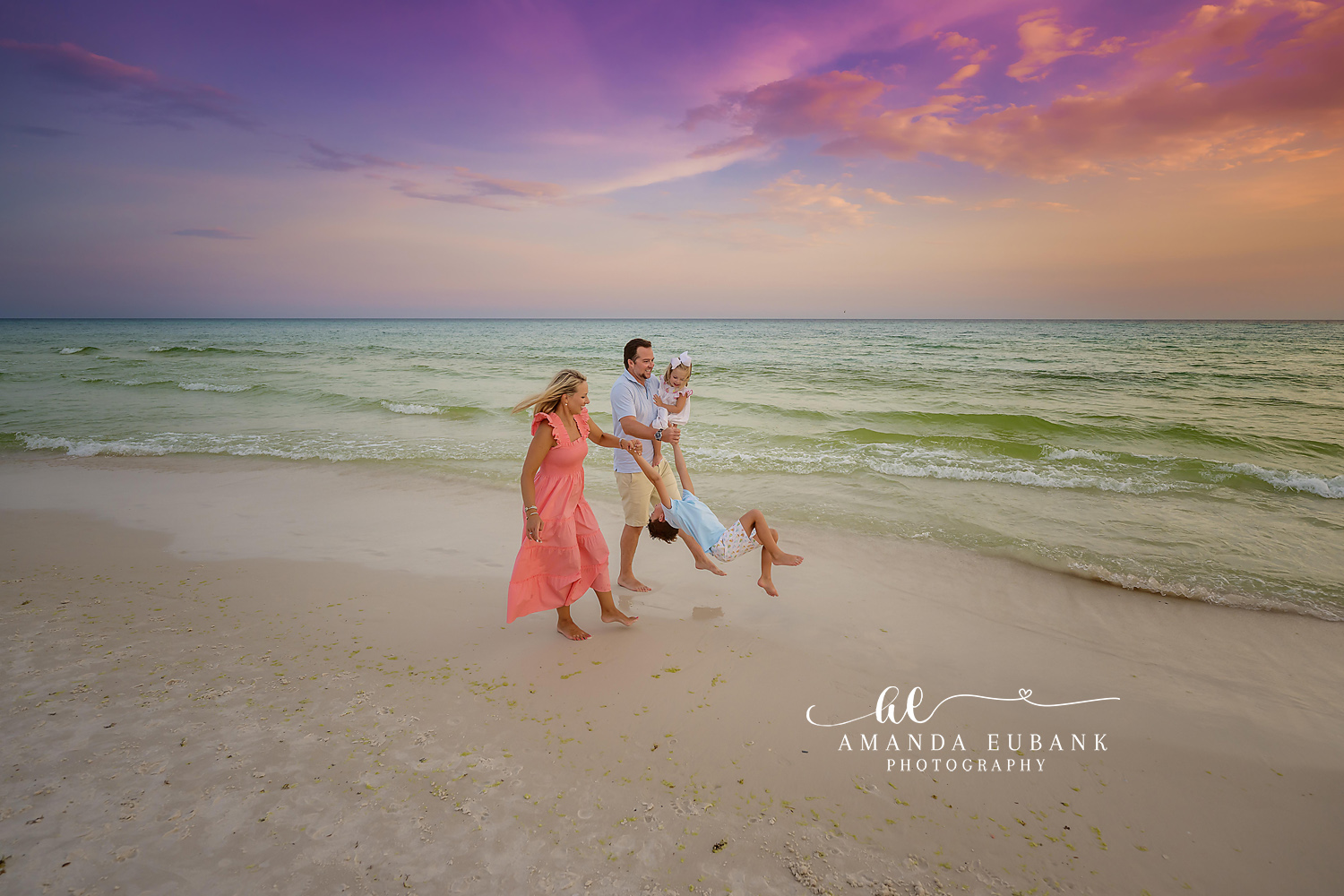 Playful family of four walking along the shoreline at dusk as the parents swing a child between them while the father carries a baby, photographed by a Santa Rosa Beach family photographer