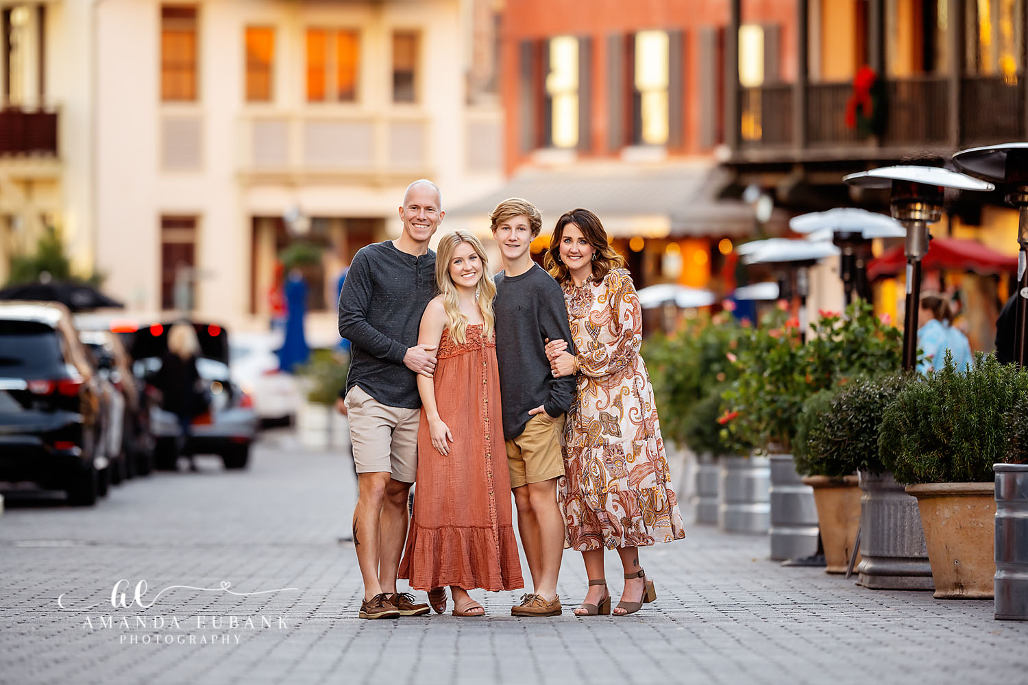 Family of four with teenage children in coordinated earth-tone outfits posing together in a seaside town square lined with shops, photographed by a Santa Rosa Beach family photographer