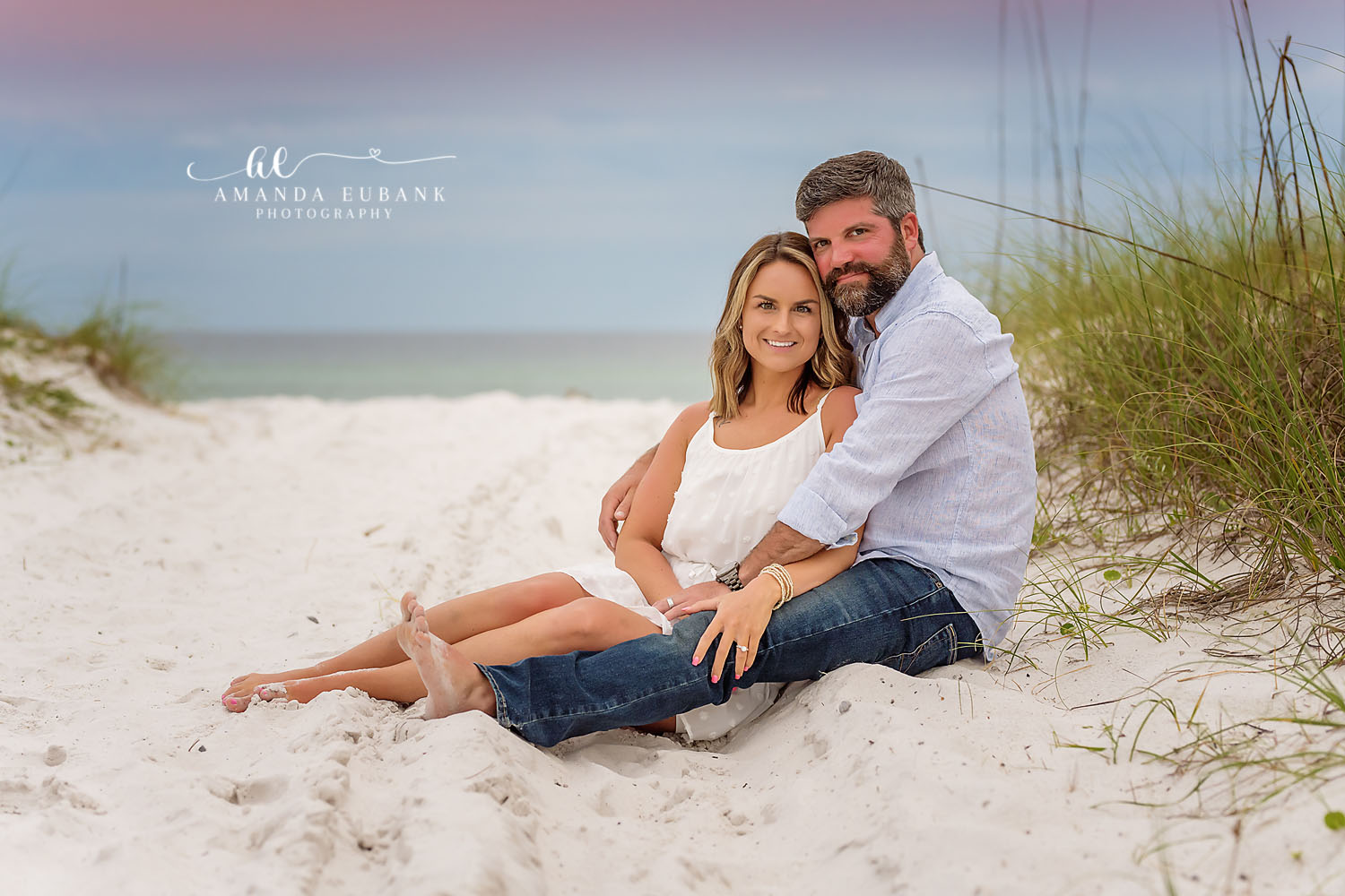 Couple cuddled together on the sand near the dunes, the woman in a white dress leaning back into her partner, photographed by a Santa Rosa Beach engagement photographer