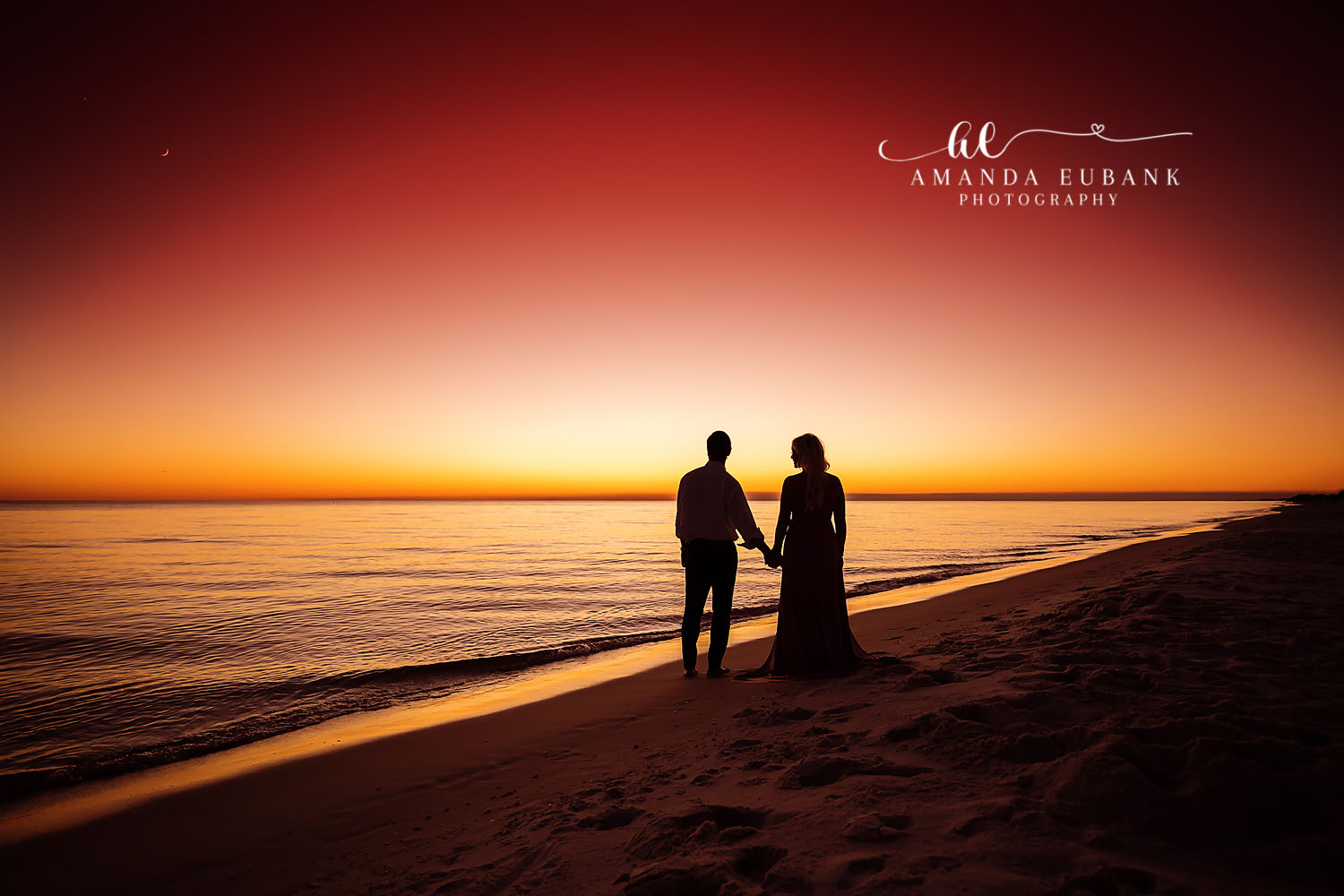 Silhouette of a couple holding hands at the shoreline against a vivid orange sunset over the gulf, photographed by a Santa Rosa Beach engagement photographer