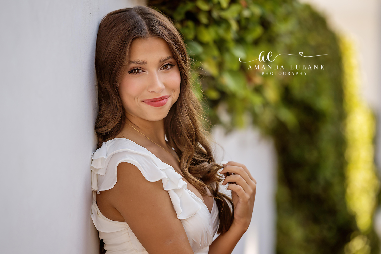 Close-up senior portrait of a young woman in a white ruffled top leaning against a wall beside soft greenery, photographed by a Santa Rosa Beach senior photographer