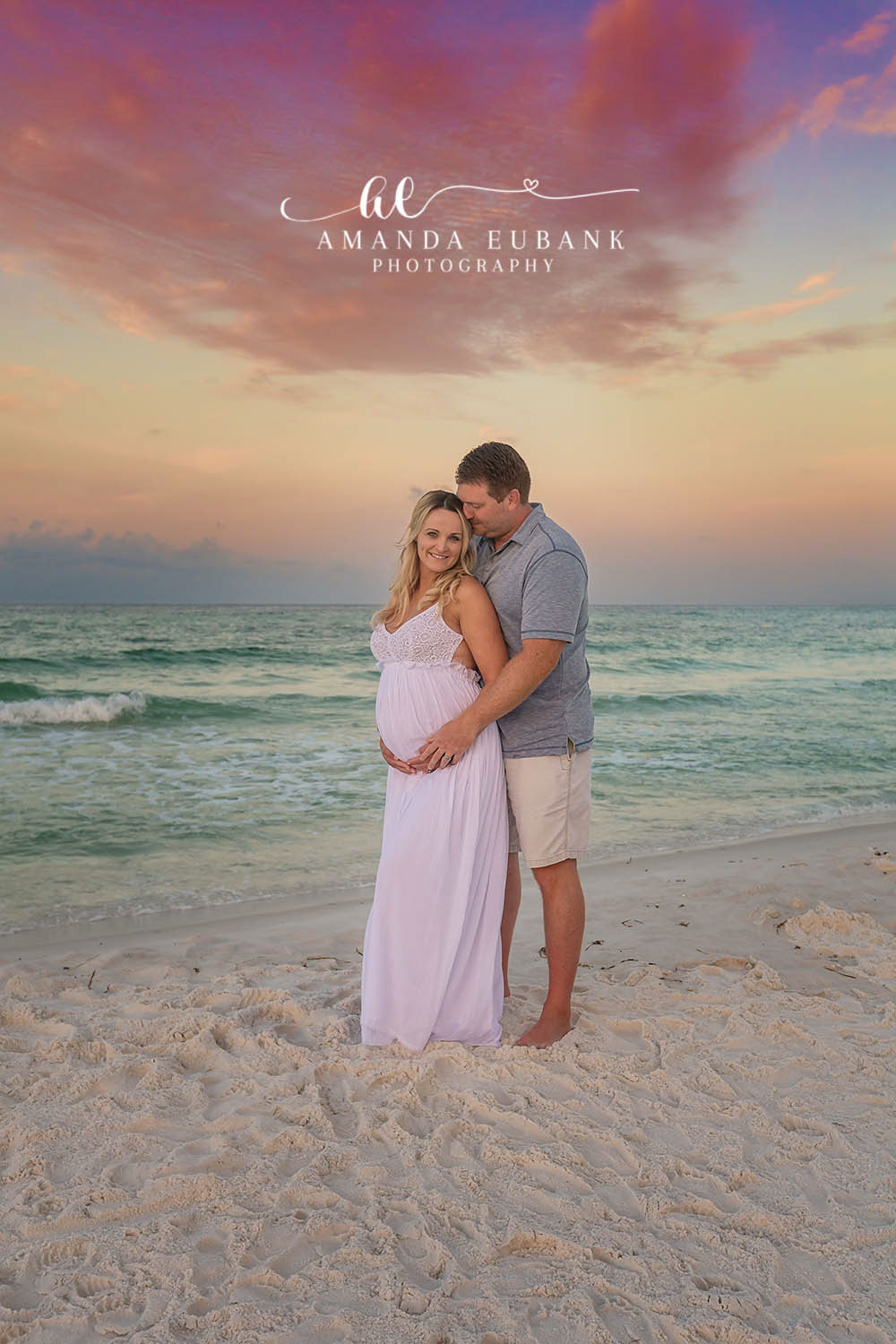 Santa Rosa Beach Maternity Photographer – Pastel Sunset Beach Maternity Session Expecting couple embracing on the beach at a pastel sunset, the woman in a flowing lavender maternity gown as both cradle her belly, photographed by a Santa Rosa Beach maternity photographer
