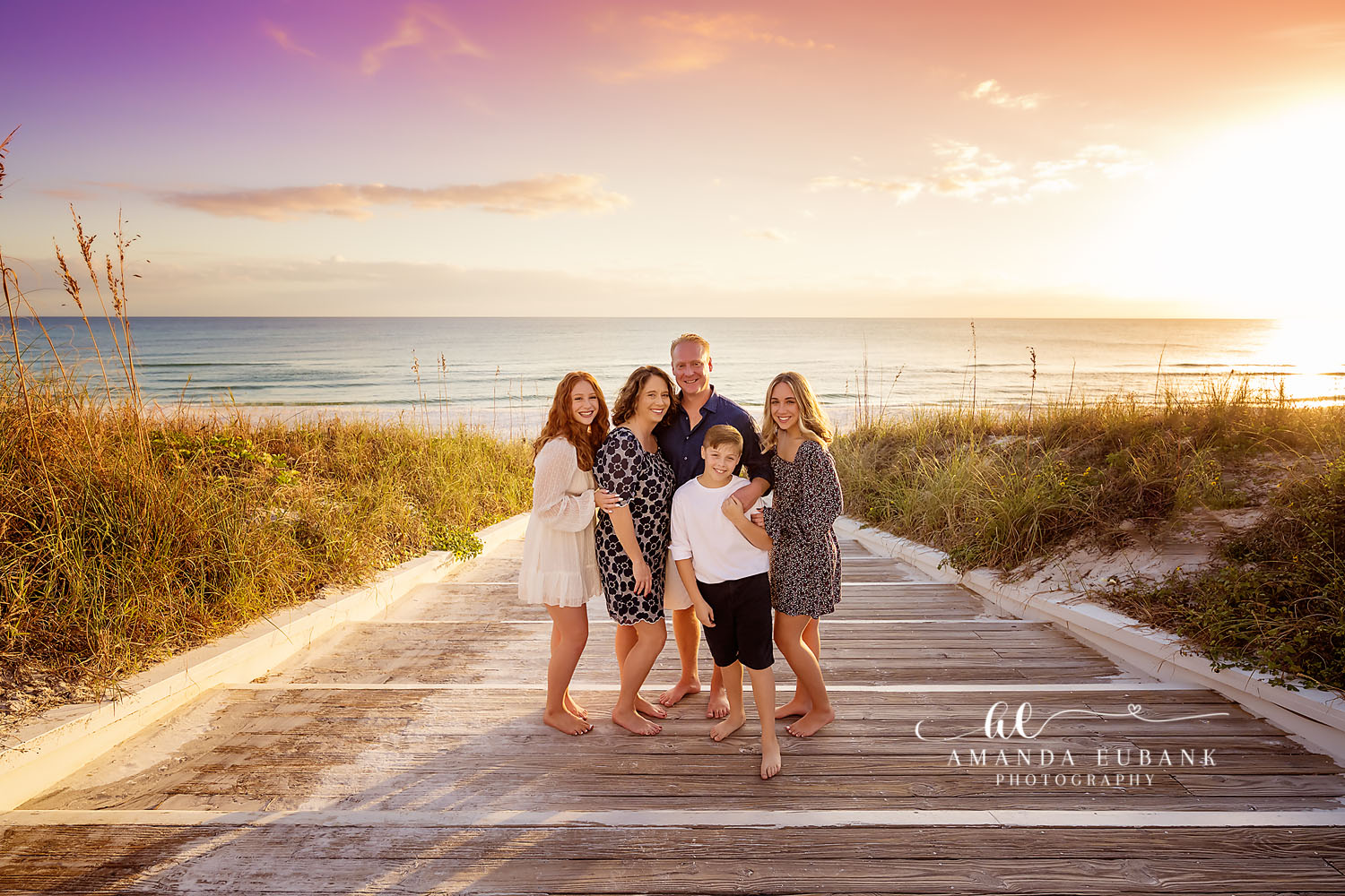 Family of five in coordinated navy and white outfits standing together on a beach boardwalk through the dunes at sunset, photographed by a Santa Rosa Beach family photographer