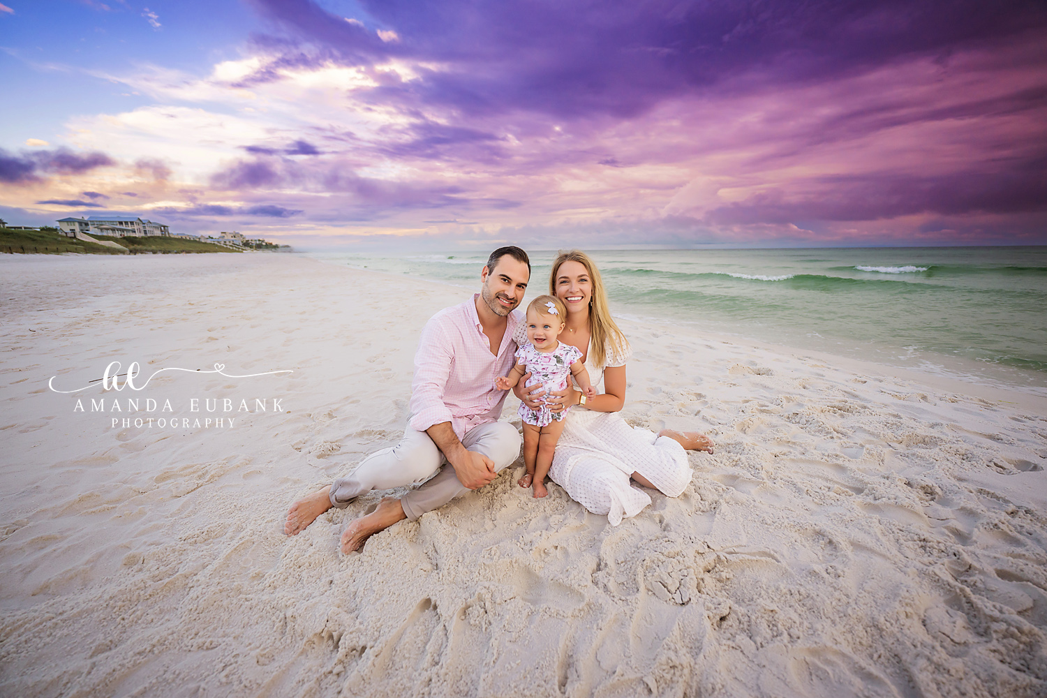 Family of three sitting together on the sand at the beach at dusk, parents with their baby daughter in coordinated light outfits, photographed by a Santa Rosa Beach family photographer