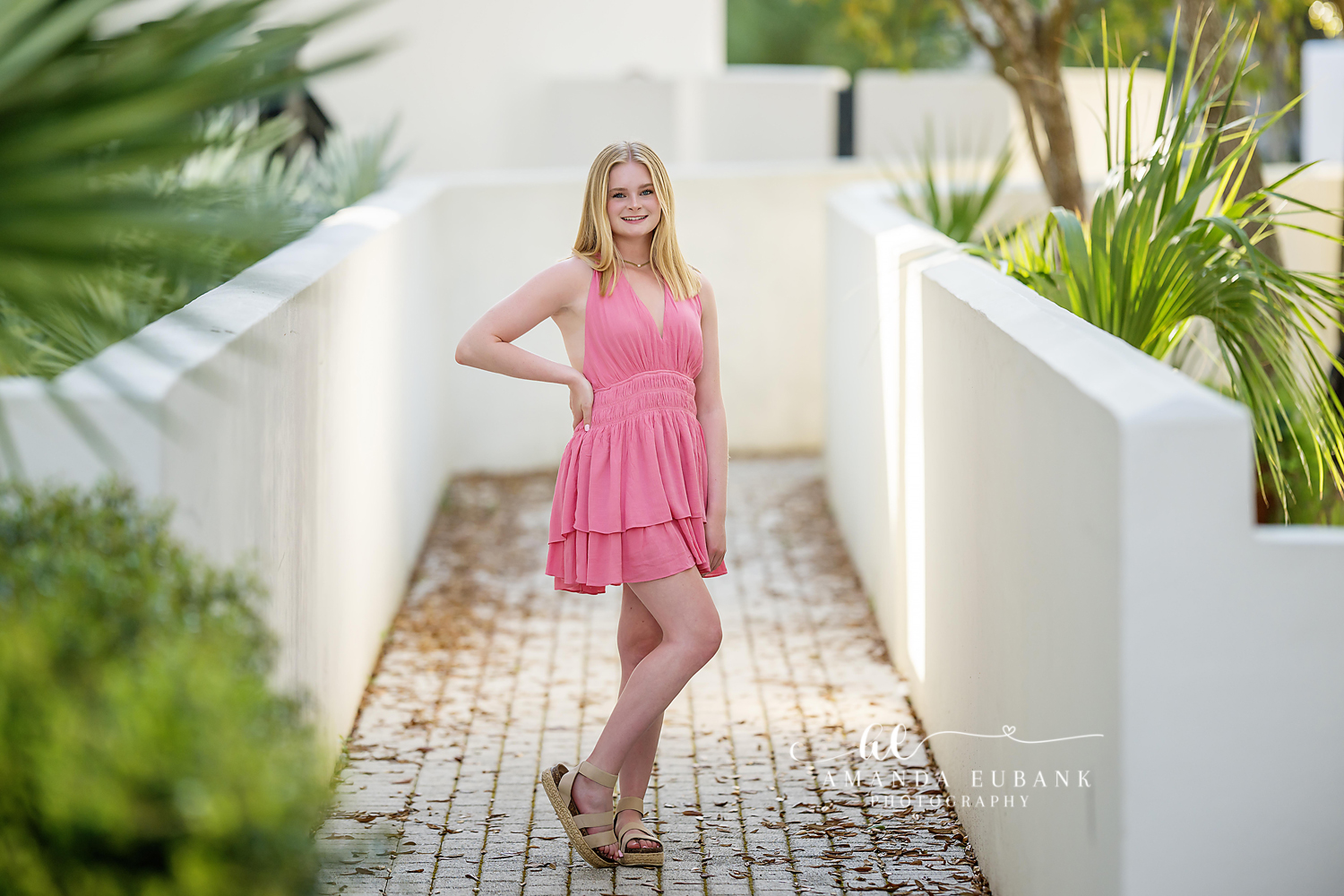 Senior girl in a pink ruffled dress posed on a brick walkway between white walls in a seaside village, photographed by a Santa Rosa Beach senior photographer