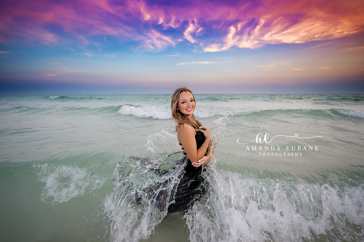 Senior girl in a flowing black gown kneeling in the breaking surf as a wave splashes around her at sunset, photographed by a Santa Rosa Beach senior photographer